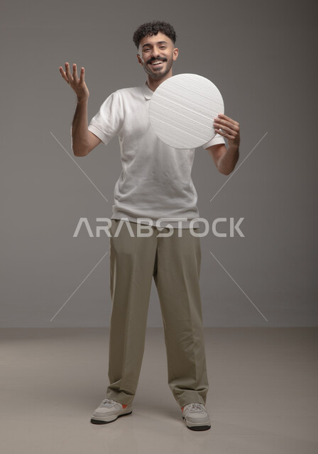 لوحه بيضاء دائريه، النظر الى الكاميرا بإيماءات السرور و الرضا، موك اب دائري أبيض، بورتريه لرجل عربي خليجي سعودي مبتسم مسرور يرتدي الزي الكاجوال و يحمل بيده لوح إعلاني فارغ، الوقوف باستقامه وثقه، صورة الجسم بالطول الكامل، خلفية رماديه