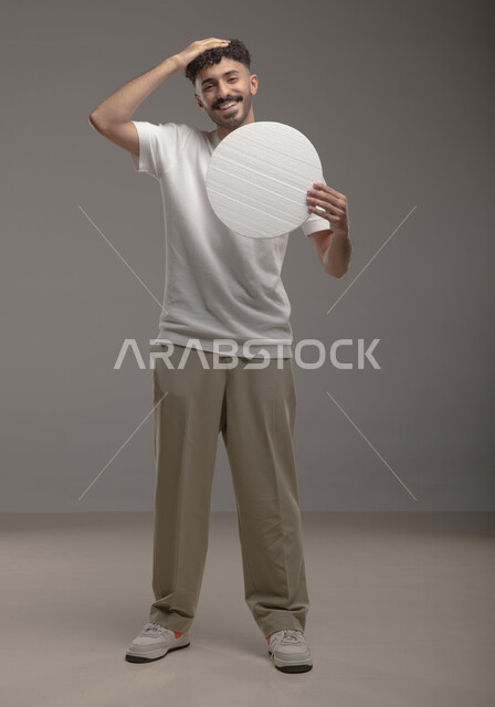 لوحه بيضاء دائريه، النظر الى الكاميرا بإيماءات السرور و الرضا، موك اب دائري أبيض، بورتريه لرجل عربي خليجي سعودي مبتسم يرتدي الزي الكاجوال و يحمل بيده لوح إعلاني فارغ، الوقوف باستقامه وثقه، صورة الجسم بالطول الكامل، خلفية رماديه