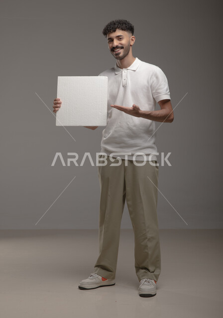 صورة الجسم بالطول الكامل، ايماءات تدل على السرور والفرح ، موك اب مربع أبيض، لوح إعلاني فارغ، بورتريه لرجل عربي خليجي سعودي مبتسم يرتدي الزي الكاجوال و يشير بيده إلى لوحه بيضاء مربعه وينظر إلى الكاميرا، الوقوف باستقامه وثقه، خلفية رماديه
