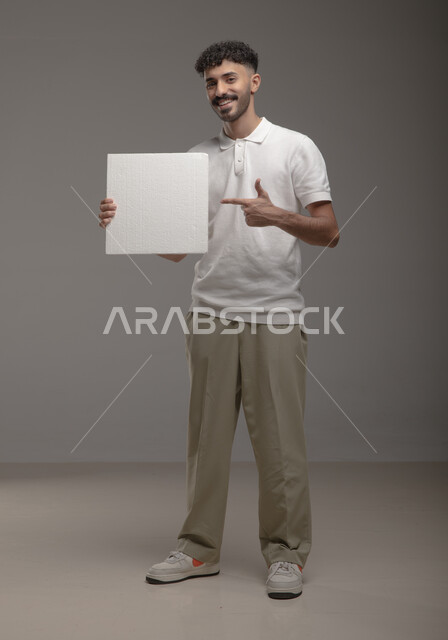 ايماءات تدل على الفرح و السرور ، موك اب مربع أبيض، لوح إعلاني فارغ، بورتريه لرجل عربي خليجي سعودي مبتسم يرتدي الزي الكاجوال و يشير باصبع السبابة إلى لوحه بيضاء مربعه وينظر إلى الكاميرا، الوقوف باستقامه وثقه، صورة الجسم بالطول الكامل، خلفية رماديه