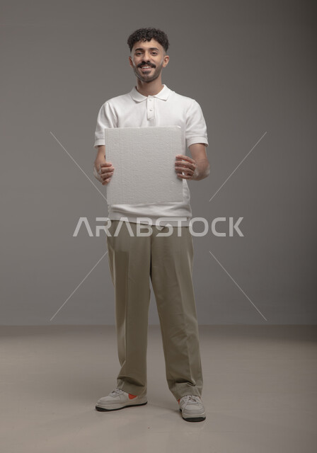 الوقوف باستقامه وثقه، لوح إعلاني فارغ، موك اب مربع أبيض، بورتريه لرجل عربي خليجي سعودي مبتسم يرتدي الزي الكاجوال و يحمل بيده لوحه بيضاء مربعه، صورة الجسم بالطول الكامل، النظر الى الكاميرا بايماءات الإعجاب والسرور، خلفية رماديه