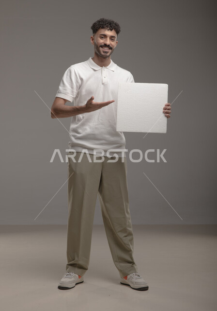 ايماءات تدل على السرور والفرح ، موك اب مربع أبيض، لوح إعلاني فارغ، بورتريه لرجل عربي خليجي سعودي مبتسم يرتدي الزي الكاجوال و يشير بيده إلى لوحه بيضاء مربعه وينظر إلى الكاميرا، الوقوف باستقامه وثقه، صورة الجسم بالطول الكامل، خلفية رماديه