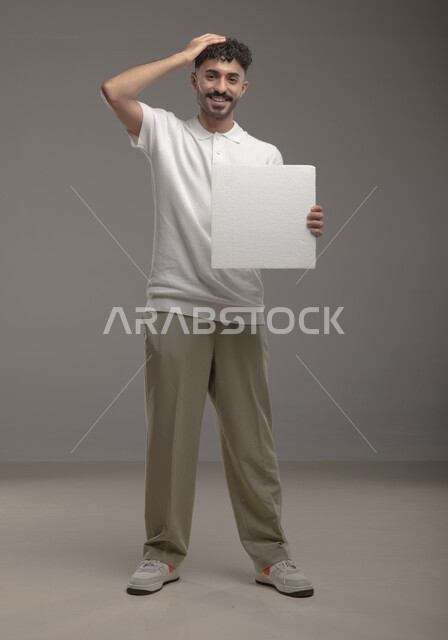 Empty billboard, looking at the camera with gestures of pleasure and satisfaction, white square mock-up, portrait of a smiling Saudi Arabian Gulf man wearing casual clothes and holding a square white board in his hand, standing upright and confident, full-length body portrait, gray background