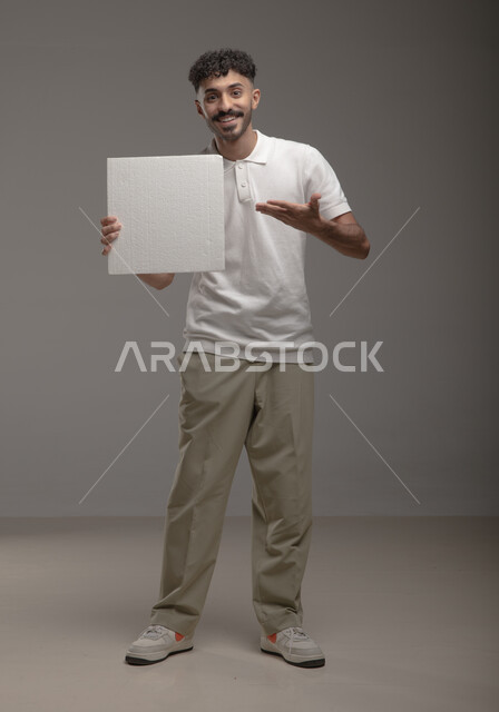 النظر الى الكاميرا بتعابير الرضا والسرور، موك اب مربع أبيض، لوح إعلاني فارغ، بورتريه لرجل عربي خليجي سعودي مبتسم يرتدي الزي الكاجوال و يشير بيده إلى لوحه بيضاء مربعه، الوقوف باستقامه وثقه، صورة الجسم بالطول الكامل، خلفية رماديه