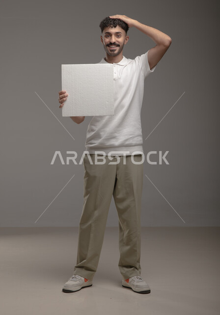  النظر الى الكاميرا بإيماءات السرور و الرضا، موك اب مربع أبيض، لوح إعلاني فارغ، بورتريه لرجل عربي خليجي سعودي مبتسم يرتدي الزي الكاجوال و يحمل بيده لوحه بيضاء مربعه، الوقوف باستقامه وثقه، صورة الجسم بالطول الكامل، خلفية رماديه