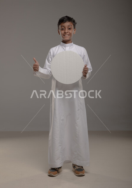 صورة الجسم بالطول الكامل، رفع الابهام الى الاعلى بإيماءات الاعجاب والرضا والنظر إلى الكاميرا ،الوقوف باستقامة وثقة، موك اب دائري ابيض، بورتريه لفتى عربي خليجي سعودي يرتدي الثوب التقليدي يحمل بيده لوحة بيضاء مدورة، لوح اعلاني فارغ،  خلفية رمادية