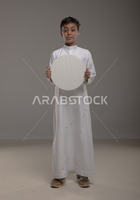 الوقوف باستقامه وثقة، لوحه بيضاء مدوره ، النظر الى الكاميرا بإيماءات السرور والاعجاب، موك اب دائري أبيض، بورتريه لفتى عربي خليجي سعودي يرتدي الزي التقليدي و يحمل بيده  لوح إعلاني فارغ، صورة الجسم بالطول الكامل، خلفية رماديه