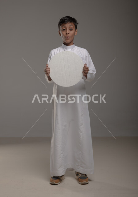 النظر الى الكاميرا بإيماءات المفاجأة والصدمة، لوحه بيضاء مدوره ،الوقوف بثقه واستقامه، موك اب دائري أبيض، بورتريه لفتى عربي خليجي سعودي يرتدي الزي التقليدي و يحمل بيده  لوح إعلاني فارغ، صورة الجسم بالطول الكامل، خلفية رماديه
