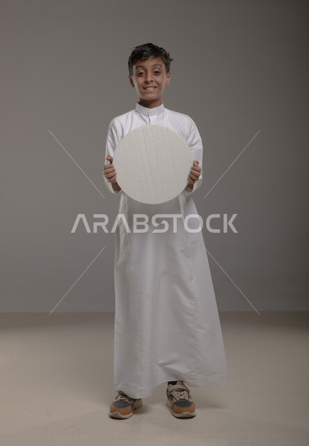 لوحه بيضاء مدوره ،الوقوف بثقه واستقامه، النظر الى الكاميرا بإيماءات السرور والاعجاب، موك اب دائري أبيض، بورتريه لفتى عربي خليجي سعودي مبتسم يرتدي الزي التقليدي و يحمل بيده  لوح إعلاني فارغ، صورة الجسم بالطول الكامل، خلفية رماديه