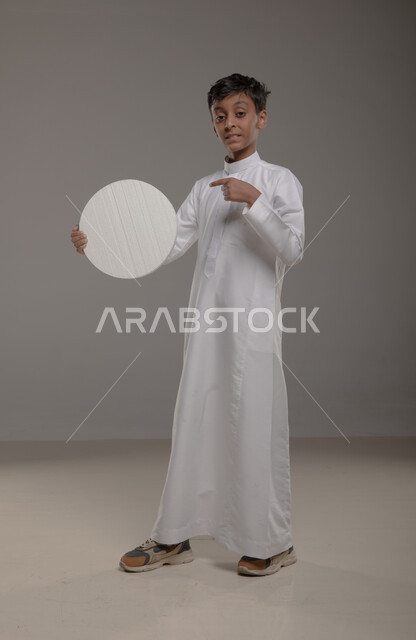 الوقوف بثقه واستقامه و النظر الى الكاميرا، لوح إعلاني فارغ، موك اب دائري أبيض، بورتريه لفتى عربي خليجي سعودي يرتدي الزي التقليدي و يشير باصبع السبابه إلى لوحه بيضاء مدوره، صورة الجسم بالطول الكامل، خلفية رماديه