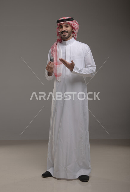 مفهوم القياده في المملكه العربيه السعوديه, الحصول على رخصة القياده, امتلاك سياره جديده بإيماءات السرور والسعاده, بورتريه لرجل عربي خليجي سعودي مبتسم يرتدي الثوب والشماغ التقليدي يشير بيده إلى مفتاح لسيارة, صورة الجسم بالطول الكامل, خلفيه رماديه