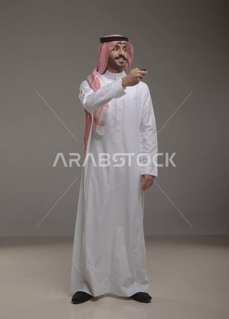 امتلاك سيارة جديده, أهميه الحصول على رخصة القياده, بورتريه لشاب عربي خليجي سعودي يرتدي الثوب والشماغ التقليدي يمسك جهاز التحكم للسيارة بإيماءات السرور, القيام بتشغيل السيارة عن بعد, صورة الجسم بالطول الكامل, خلفيه رماديه