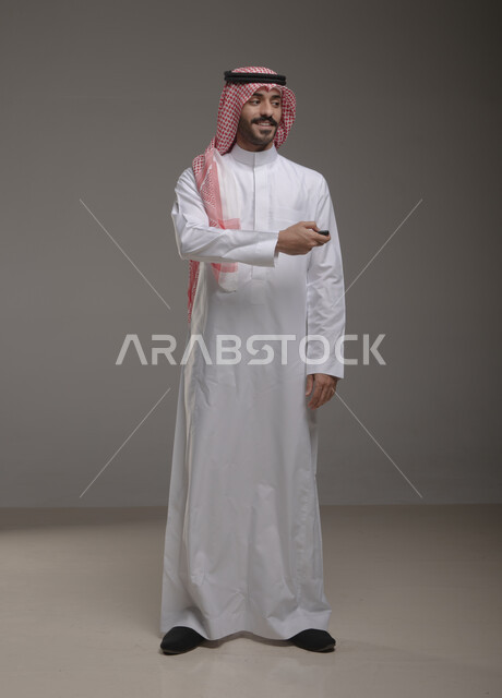 القيام بتشغيل السيارة عن بعد, أهميه الحصول على رخصة القياده, بورتريه لشاب عربي خليجي سعودي يرتدي الثوب والشماغ التقليدي يمسك جهاز التحكم للسيارة بإيماءات السرور, امتلاك سيارة جديده, صورة الجسم بالطول الكامل, خلفيه رماديه