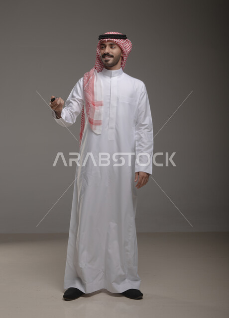 أهميه الحصول على رخصة القياده, بورتريه لشاب عربي خليجي سعودي يرتدي الثوب والشماغ التقليدي يمسك جهاز التحكم للسيارة بإيماءات السرور, القيام بتشغيل السيارة عن بعد, امتلاك سيارة جديده, صورة الجسم بالطول الكامل, خلفيه رماديه