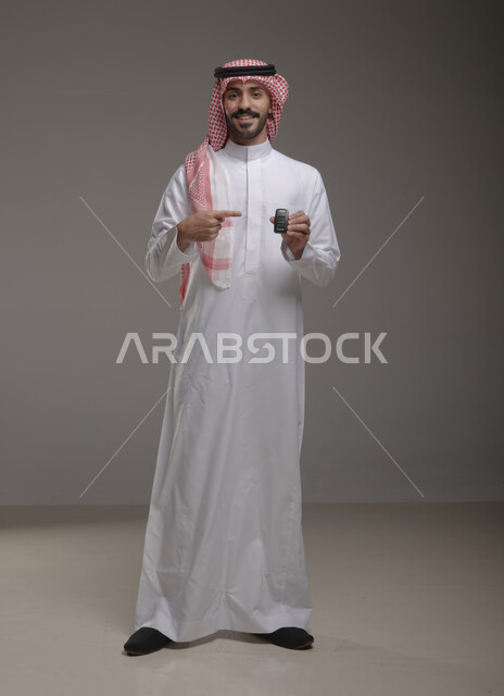 The concept of driving in Saudi Arabia, expressing feelings of joy and joy in owning a new car, portrait of a Saudi Gulf Arab man wearing the traditional thobe and shemagh holding the remote control key, pointing to the key with happy gestures, full-length body portrait, gray background