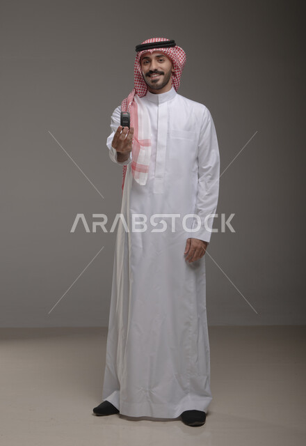 استلام وتسليم المفاتيح, ايماءات السعاده والسرور بامتلاك سياره جديده, بورتريه لرجل عربي خليجي سعودي يرتدي الثوب والشماغ التقليدي يمسك مفتاح السيارة بيده وينظر إلى الكاميرا, الحصول على خدمه صف السيارات بالمواقف الخاصه, صورة الجسم بالطول الكامل, خلفيه رماديه
