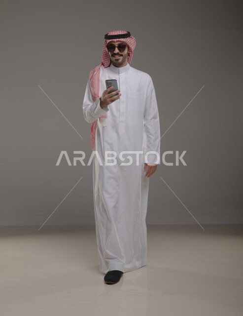 التسوق الالكتروني الذكي والأمن عبر الانترنت, بورتريه لرجل عربي خليجي سعودي مبتسم يرتدي النظاره الشمسيه والثوب والشماغ التقليدي يقوم باستخدام الهاتف المحمول, تصفح مواقع التواصل الاجتماعي بإيماءات السرور والسعاده, صورة الجسم بالطول الكامل, خلفيه رماديه