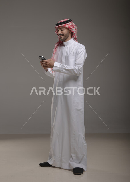 Using advanced modern technologies, chatting and friendly conversations with friends via the Internet, a portrait of a smiling Saudi Arabian Gulf man wearing a thobe and shemagh browsing social networking sites via mobile phone with focus gestures, full-length body image, gray background