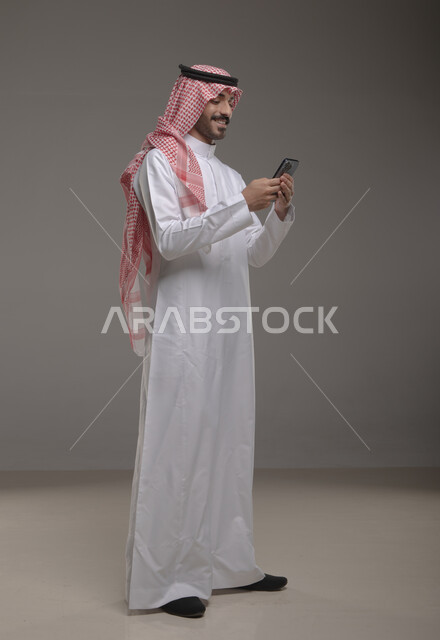 الدردشه والمحادثات الوديه مع الأصدقاء عبر الانترنت, بورتريه لشاب عربي سعودي خليجي مبتسم يرتدي الثوب والشماغ يقوم بتصفح مواقع التواصل الاجتماعي عبر الهاتف المحمول بإيماءات التركيز, استخدام التقنيات الحديثة المتطورة, صورة الجسم بالطول الكامل, خلفيه رماديه