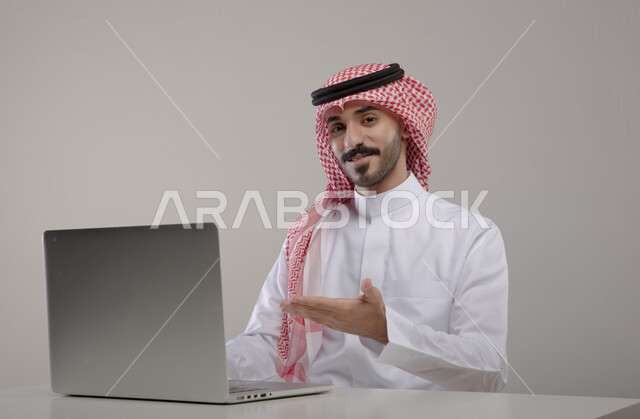 التسويق والترويج الإعلاني, العمل في بيئة مكتبيه مريحه, انجاز مهام العمل عبر الأونلاين, بورتريه لشاب عربي خليجي سعودي مبتسم يرتدي الشماغ والثوب التقليدي ينظر إلى الكاميرا ويشير بيده إلى جهاز الحاسوب بإيماءات الإعجاب, خلفيه رماديه