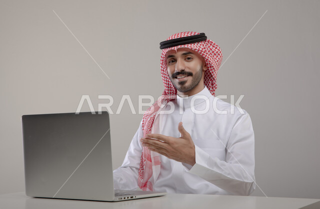 الترويج والتسويق الإعلاني, العمل في بيئة مكتبيه مريحه, انجاز مهام العمل عبر الأونلاين, بورتريه لشاب عربي خليجي سعودي مبتسم يرتدي الشماغ والثوب التقليدي ينظر إلى الكاميرا ويشير بيده إلى جهاز الحاسوب بإيماءات الإعجاب, خلفيه رماديه