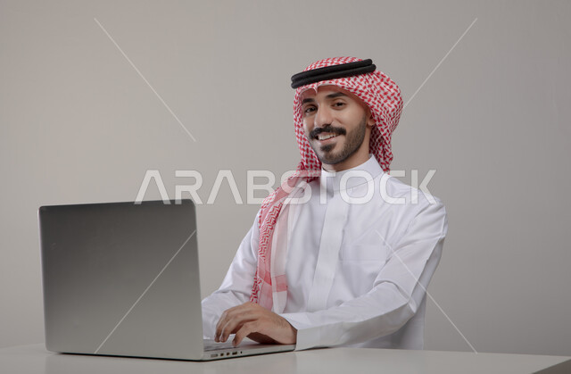 Completing work and tasks remotely, spending enjoyable times browsing social media, portrait of a smiling Saudi Gulf Arab young man wearing a shemagh and traditional dress looking at the camera with gestures of pleasure and happiness, conducting job interviews online, gray background