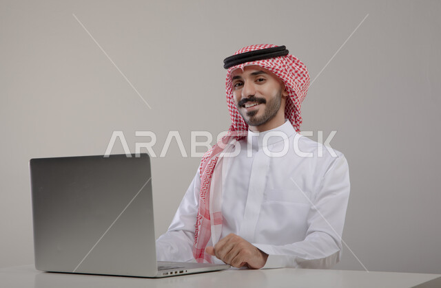 قضاء أوقات ممتعه بتصفح وسائل التواصل الاجتماعي, انجاز الأعمال والمهام عن بعد, بورتريه لشاب عربي خليجي سعودي مبتسم يرتدي الشماغ والثوب التقليدي ينظر إلى الكاميرا بإيماءات السرور والسعاده, إجراء مقابلات عمل أونلاين, خلفيه رماديه