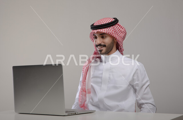 Conducting job interviews online, spending enjoyable times browsing social media, completing work and tasks remotely, a portrait of a smiling Saudi Gulf Arab young man wearing a shemagh and traditional thobe, looking at the computer with gestures of pleasure and happiness, gray background.