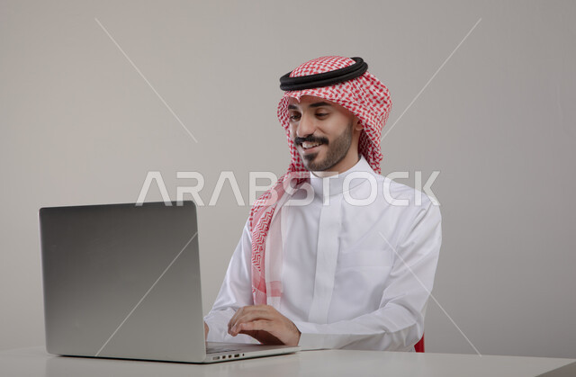 استخدام الحاسب المحمول في الأعمال المكتبيه, تصفح الانترنت والعمل عن بعد عبر جهاز الابتوب, بورتريه مقرب لشاب عربي خليجي سعودي مبتسم يرتدي الثوب والشماغ التقليدي يجلس في مكتبه, التسويق الالكتروني الذكي والآمن عبر وسائل السوشال ميديا, خلفيه رماديه