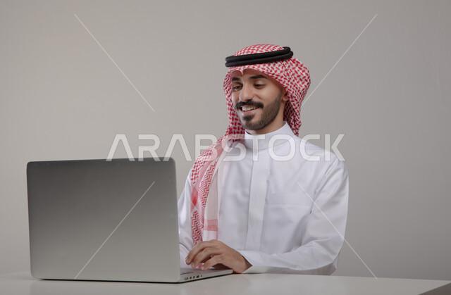 تصفح الانترنت والعمل عن بعد عبر جهاز الابتوب, بورتريه مقرب لشاب عربي خليجي سعودي مبتسم يرتدي الثوب والشماغ التقليدي يجلس في مكتبه, استخدام الحاسب المحمول في الأعمال المكتبيه, التسويق الالكتروني الذكي والآمن عبر وسائل السوشال ميديا, خلفيه رماديه