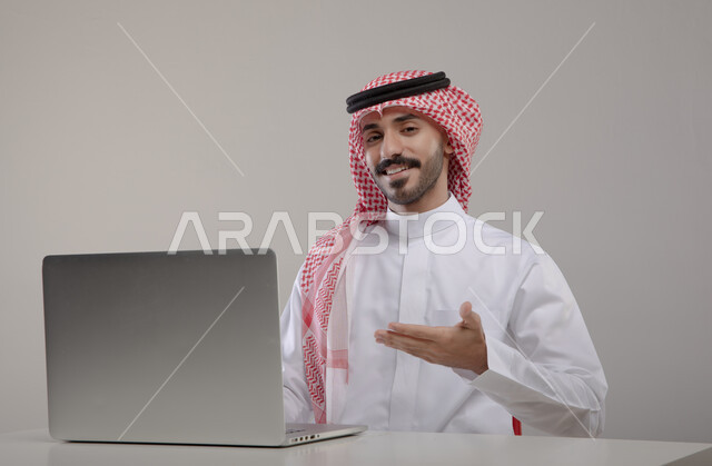 العمل في بيئة مكتبيه مريحه, انجاز مهام العمل عبر الأونلاين, بورتريه لشاب عربي خليجي سعودي مبتسم يرتدي الشماغ والثوب التقليدي ينظر إلى الكاميرا ويشير بيده إلى جهاز الحاسوب بإيماءات الإعجاب, الترويج والتسويق الإعلاني, خلفيه رماديه