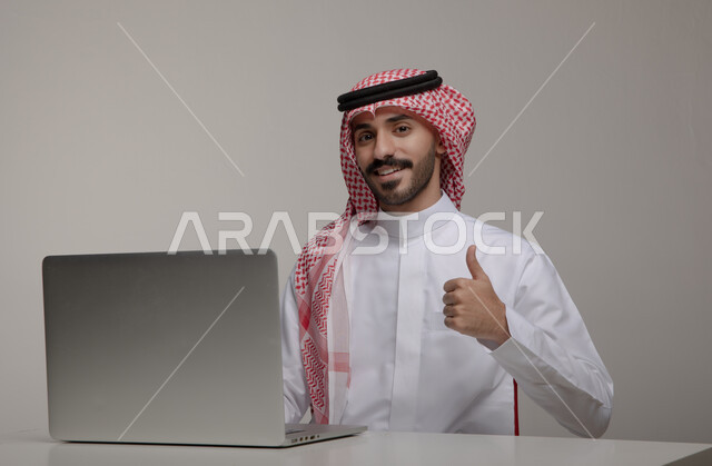 Completing work tasks via the laptop, the concept of integrating work with technology and technology, a portrait of a Saudi Gulf Arab man wearing the traditional thobe and shemagh, raising his finger with gestures of approval and admiration, using electronic applications in Saudi offices and jobs, gray background