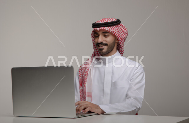 التسويق الالكتروني الذكي والآمن عبر وسائل السوشال ميديا, تصفح الانترنت والعمل عن بعد عبر جهاز الابتوب, بورتريه مقرب لشاب عربي خليجي سعودي مبتسم يرتدي الثوب والشماغ التقليدي يجلس في مكتبه, استخدام الحاسب المحمول في الأعمال المكتبيه, خلفيه رماديه