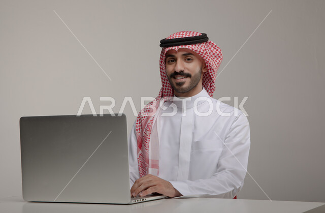 The concept of integrating work with technology and technology, completing work tasks via a laptop, a portrait of a smiling Saudi Gulf Arab man wearing the traditional thobe and shemagh looking at the camera with gestures of pleasure, using electronic applications in Saudi offices and jobs, gray background