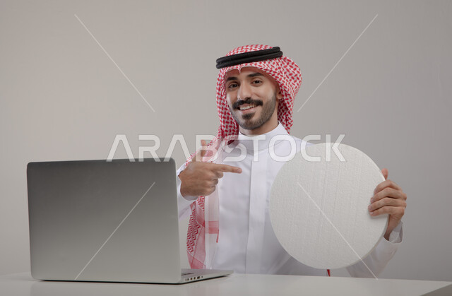 موك اب دائري ابيض, لوح اعلاني فارغ, بورتريه لشاب عربي خليجي سعودي مبتسم يرتدي الثوب والشماغ التقليدي يجلس أمام الابتوب ويشير بيده إلى لوحه بيضاء مدوره, التسويق الرقمي الالكتروني عبر السوشال ميديا, النظر إلى الكاميرا بإيماءات السرور والإعجاب, خلفيه رماديه