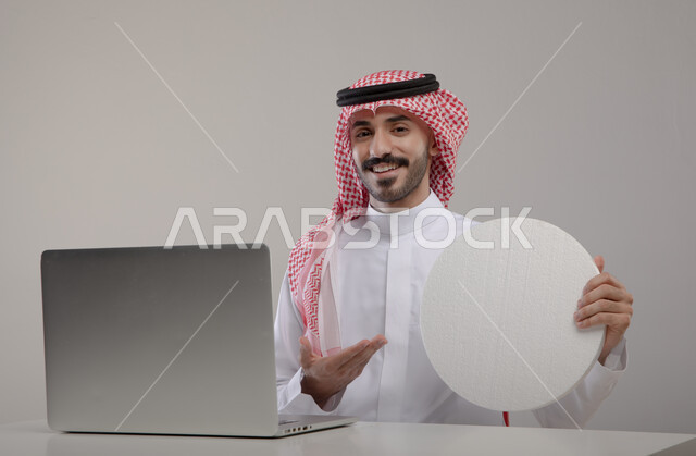 لوح اعلاني فارغ، موك اب دائري ابيض, بورتريه لشاب عربي خليجي سعودي مبتسم يرتدي الثوب والشماغ التقليدي يجلس أمام الابتوب ويشير بيده إلى لوحه بيضاء مدوره, التسويق الرقمي الالكتروني عبر السوشال ميديا, النظر إلى الكاميرا بإيماءات السرور والإعجاب, خلفيه رماديه