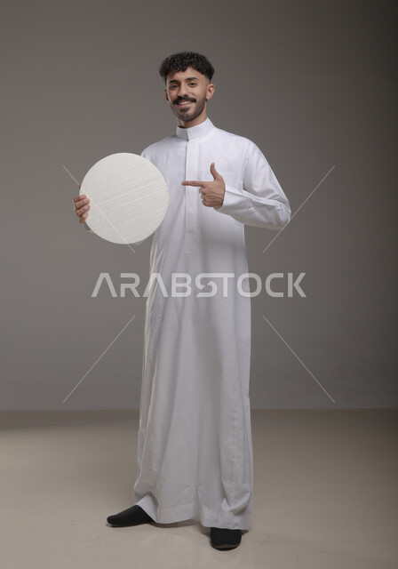 النظر الى الكاميرا بإيماءات السرور والاعجاب، لوح إعلاني فارغ، موك اب دائري أبيض، بورتريه لرجل عربي خليجي سعودي مبتسم يرتدي الزي التقليدي و يشير باصبع السبابه إلى لوحه بيضاء مدوره، الوقوف باستقامه وثقه، صورة الجسم بالطول الكامل، خلفية رماديه