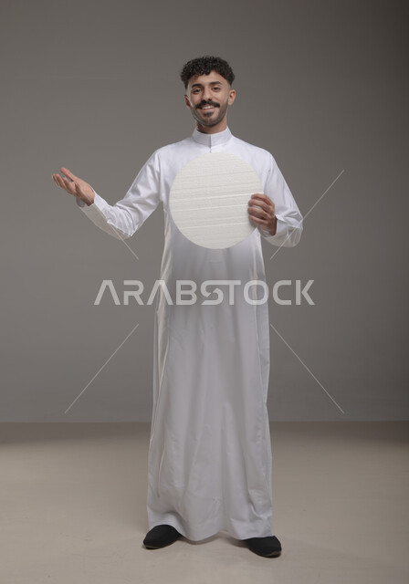 عرض لوحه بيضاء دائريه، النظر الى الكاميرا بإيماءات السرور و الرضا، موك اب دائري أبيض، بورتريه لرجل عربي خليجي سعودي مبتسم يرتدي الزي التقليدي و يحمل بيده لوح إعلاني فارغ، الوقوف باستقامه وثقه، صورة الجسم بالطول الكامل، خلفية رماديه