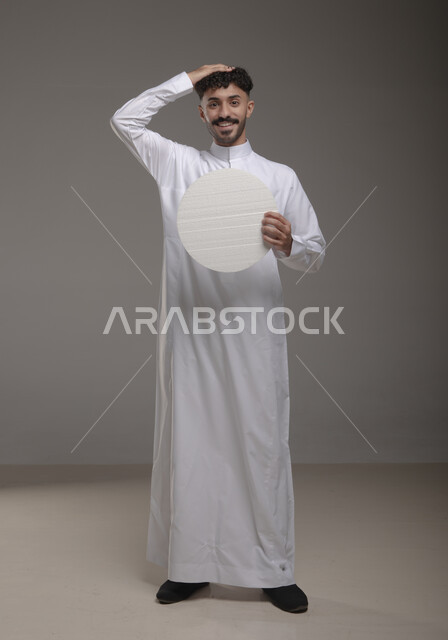 عرض لوحه بيضاء دائريه، النظر الى الكاميرا بإيماءات السرور و الرضا، موك اب دائري أبيض، بورتريه لرجل عربي خليجي سعودي مبتسم يرتدي الزي التقليدي و يحمل بيده لوح إعلاني فارغ، الوقوف باستقامه وثقه، صورة الجسم بالطول الكامل، خلفية رماديه