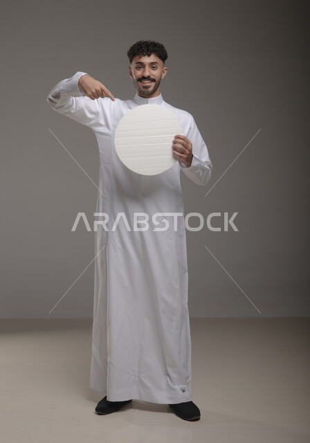 النظر الى الكاميرا بإيماءات السرور والاعجاب، لوح إعلاني فارغ، موك اب دائري أبيض، بورتريه لرجل عربي خليجي سعودي مبتسم يرتدي الزي التقليدي و يشير باصبع السبابه إلى لوحه بيضاء مدوره، الوقوف باستقامه وثقه، صورة الجسم بالطول الكامل، خلفية رماديه