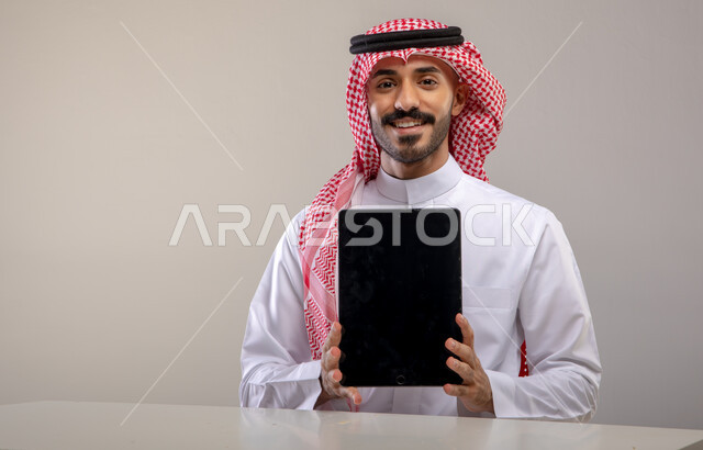 استعمال التقنيات الحديثة للتسويق الرقمي الالكتروني، عرض تطبيقات وخدمات الجهاز اللوحي، بورتريه لشاب عربي خليجي سعودي مبتسم يرتدي الشماغ و الثوب التقليدي يجلس على الطاولة وينظر الى الكاميرا و يحمل بيده التابلت بشاشة سوداء فارغة، خلفية رمادية