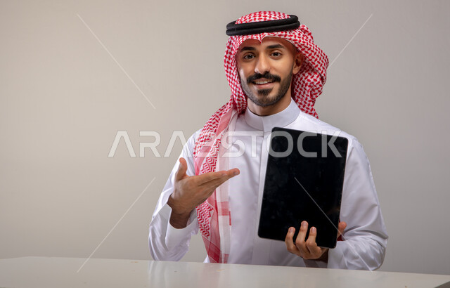 إيماءات الإشارة الى الجهاز اللوحي بشاشة سوداء فارغة، بورتريه لشاب عربي خليجي سعودي مبتسم يرتدي الشماغ و الثوب التقليدي يجلس على الطاولة وينظر الى الكاميرا و يحمل بيده التابلت، استعمال التقنيات الحديثة للتسويق الرقمي الالكتروني، خلفية رمادية