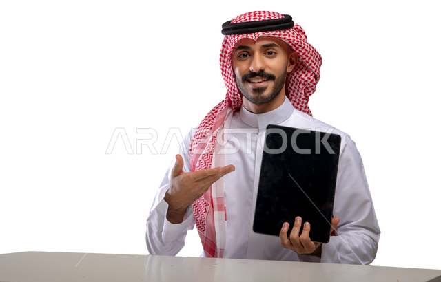 Gestures pointing to the tablet with a blank black screen, portrait of a smiling Saudi Gulf Arab young man wearing a shemagh and traditional dress, sitting