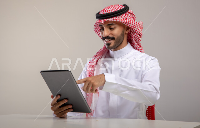 التسوق والشراء عن طريق الانترنت ، إيماءات النقر والضغط على الجهاز اللوحي, بورتريه لرجل عربي خليجي سعودي مبتسم يرتدي الشماغ والثوب التقليدي يجلس على الطاولة و يحمل بيده التابليت، الاستمتاع باستخدام برامج و تطبيقات الأجهزة اللوحية, خلفية رمادية