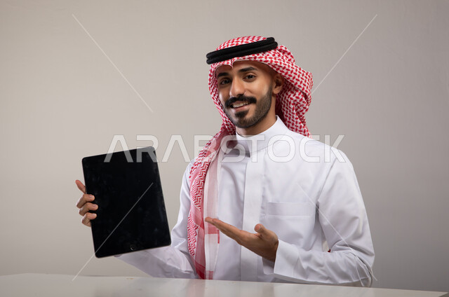 عرض تطبيقات وخدمات الجهاز اللوحي، بورتريه لشاب عربي خليجي سعودي مبتسم يرتدي الشماغ و الثوب التقليدي يجلس على الطاولة وينظر الى الكاميرا و يحمل بيده التابلت بشاشة سوداء فارغة ويشير اليه، استعمال التقنيات الحديثة للتسويق الرقمي الالكتروني، خلفية رمادية