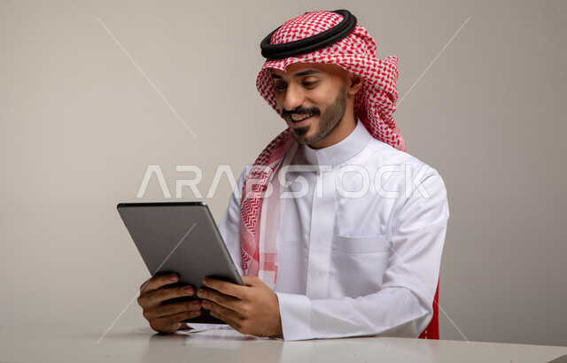 التواصل مع الأهل والأصدقاء عن بعد, بورتريه لرجل عربي خليجي سعودي مبتسم يرتدي الشماغ والثوب التقليدي يجلس على الطاولة و يحمل بيده جهاز التابليت، التسوق والشراء عن طريق الانترنت ، إيماءات الاستمتاع باستخدام برامج و تطبيقات الأجهزة اللوحية, خلفية رمادية