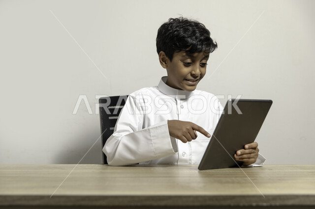 Using mobile devices for learning and entertainment, following educational and entertainment programs with happy gestures, portrait of a smiling Saudi Gulf Arab boy wearing a traditional dress sitting at the desk and tapping on the tablet, white background