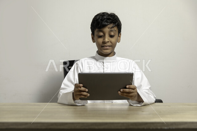 Gestures of integration and focus while watching educational and entertaining programs on the tablet, portrait of a smiling Saudi Gulf Arab boy wearing a traditional dress and holding a tablet in his hand, using mobile devices for learning and entertainment, white background