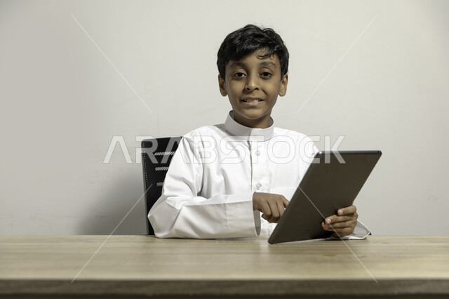 قضاء الوقت على الأجهزة المحمولة في التعلم والترفيه، متابعة البرامج التعليمية والترفيهية بإيماءات السرور، بورتريه لولد عربي خليجي سعودي مبتسم يرتدي الثوب التقليدي يجلس على المكتب ويقوم بالنظر الى الكاميرا والنقر على جهاز التابليت، خلفية بيضاء