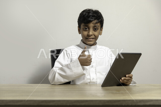 Watching educational and entertainment programs with pleasure, sitting at the table and looking at the camera with gestures indicating admiration and a sign of quality, portrait of a smiling Gulf Arab Saudi boy wearing a traditional dress and holding a tablet in his hand, using mobile devices for learning and entertainment, white background